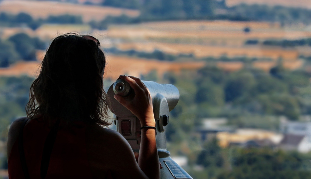 Young woman at a viewpoint looking through binoculars trying to find the best real estate agent in Colorado